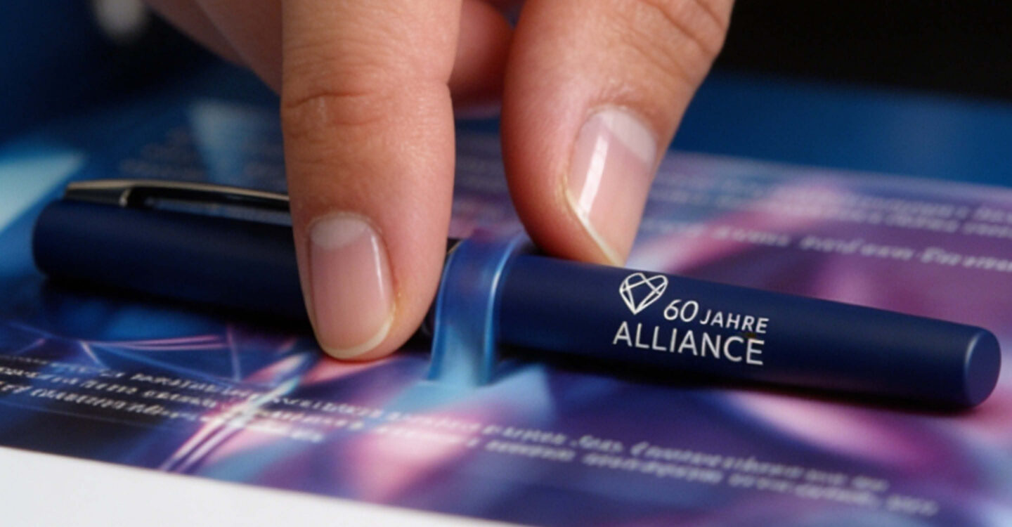 Detailaufnahme eines Kugelschreibers mit Aufdruck ‚60 Jahre Alliance‘, gehalten von einer Hand auf einem farblich leuchtenden Werbeflyer