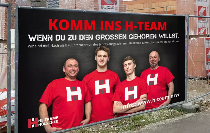Bild von einem Bauzaunbanner der Personalkampagne mit der Aufschrift "Komm ins H-Team wenn du zu den großen gehören willst. Wir sind mehrfach als Bauunternehmen des Jahres ausgezeichnet. Heitkamp & Hülscher - mehr als nur ein Job"
